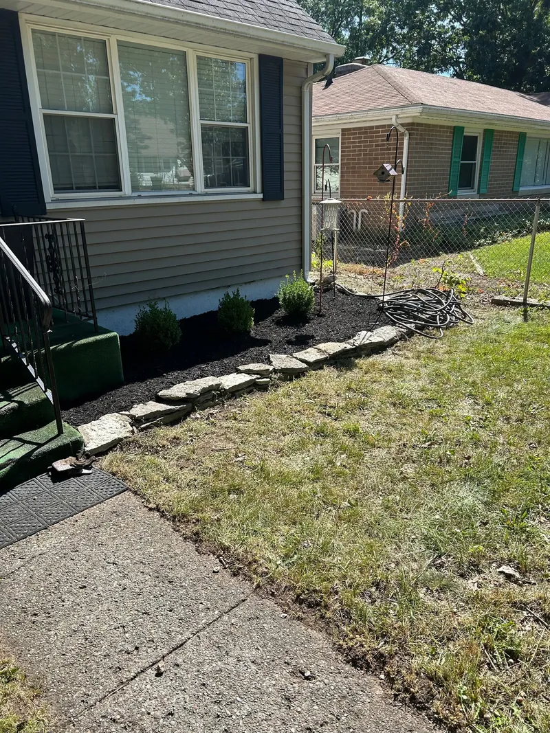 Dark mulch, small shrubs, and a stone border below a blue-shuttered window
